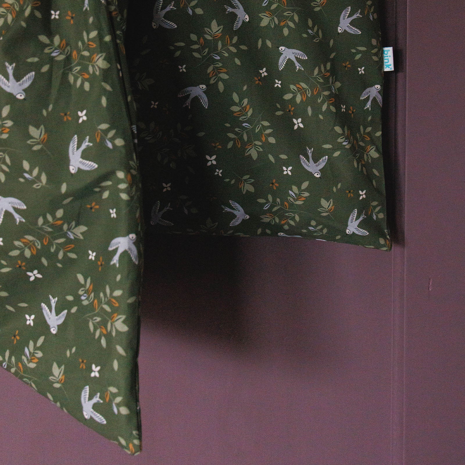 Sfeerbeeld van dekbedovertrek Merel met een mooie print met vogels, bloemen en takken met bladeren in lichtgroene tinten, caramel bloemen en lila accenten, hangend aan kast..